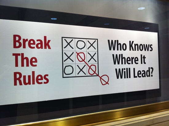 A sign that shows a tic tac toe board with an extra o outside the box giving the rule breaker the "win." Next to game squares it reads: "Break The Rules" and "Who Knows Where it Will Lead?"
