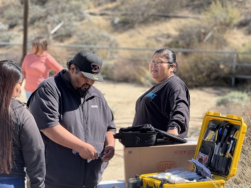Angela looks at her crew explore fiber splicing equipment on a table outdoors on RantenenTown Ranch in the desert-like foothills of Anguana, Califonia