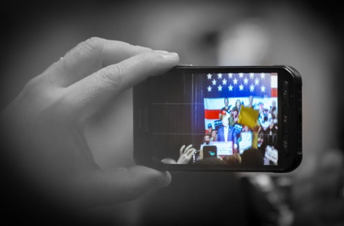 A black and white hand holds up a cell phone to photograph President Trump
