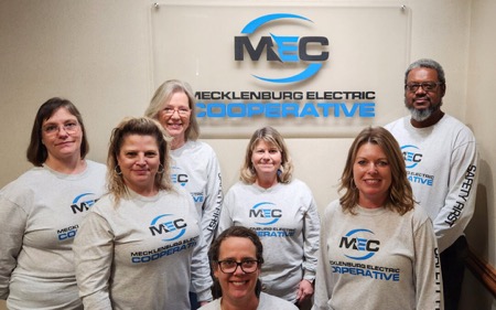 A group of seven Mecklenberg Electric Coop staff stand in front of MEC logo
