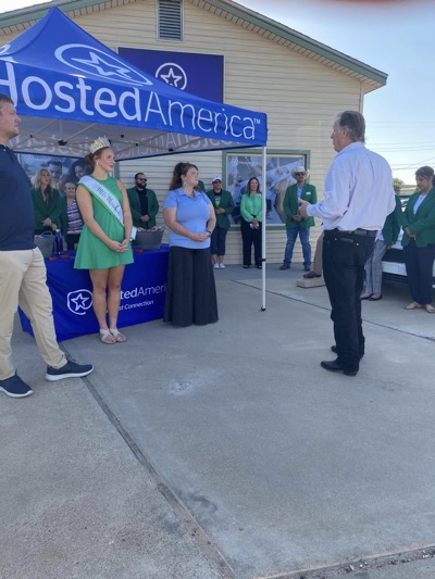 A beauty pageant contestant stands outside Hosted America tent covered table speaking to community member