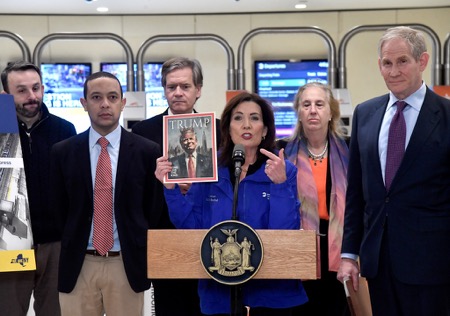 NY Gov Kathy Hochul surrounded by officials at a podium holding up a magazine with President Trump on the cover