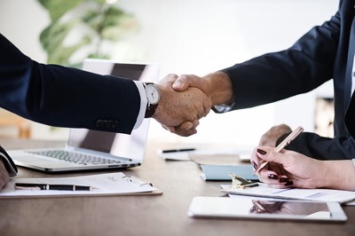 A handshake over a desk with laptop in background and woman's hand holding a pen over official agreement
