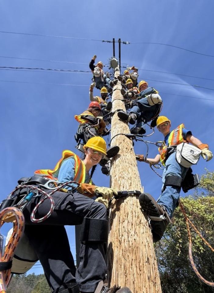 The graduating class of the Vermont fiber apprenticeship program smile down at camera while a half dozen technicians are on a utility pole