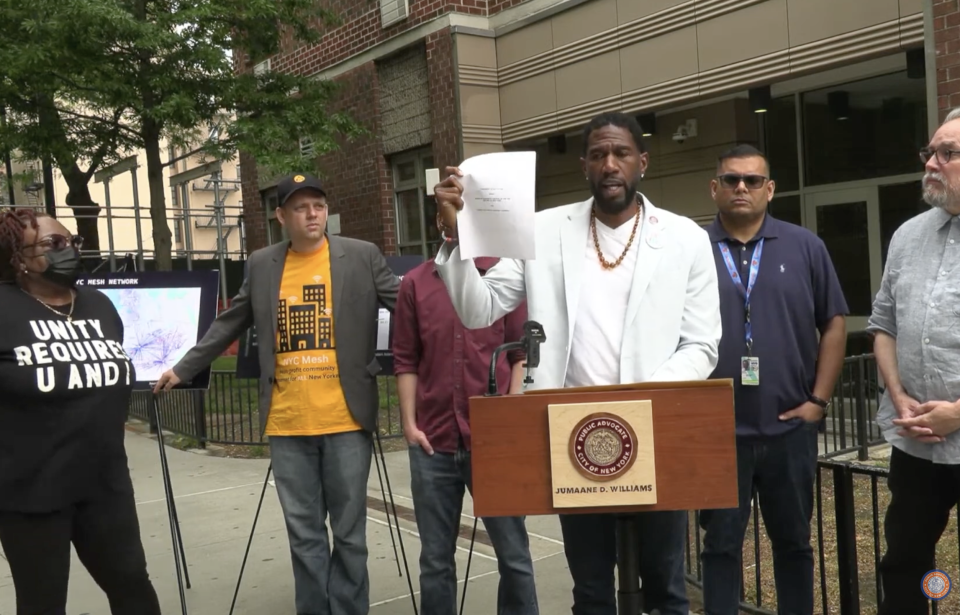NYC Public Advocate Jumaane Williams stands a podium flanked by advocates and a NYC Mesh representative in front of an apartment building