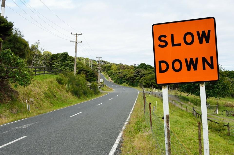 An orange sign on side of rural road reads: "Slow Down"